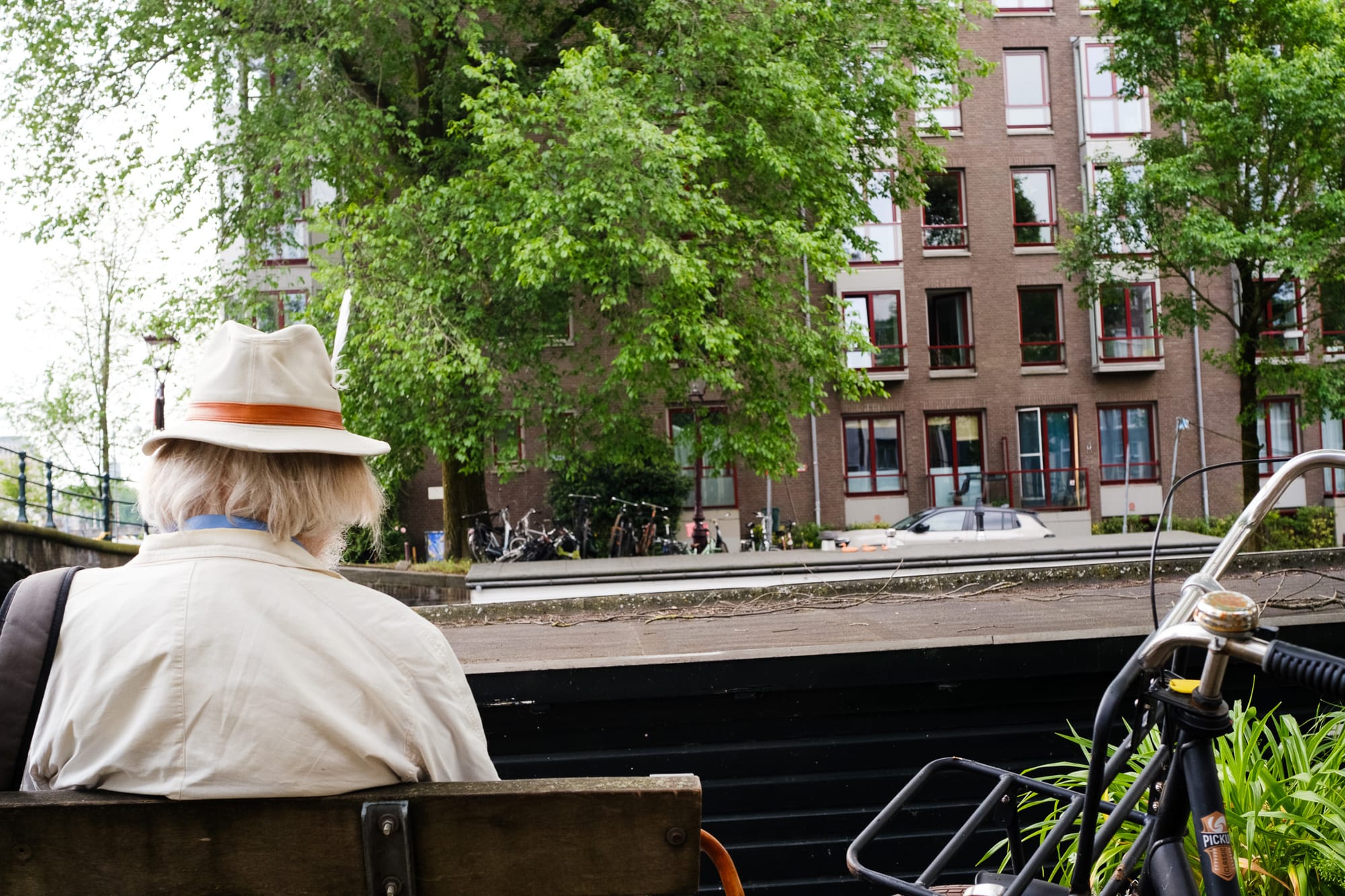 Person in a hat sitting on a bench by an Amsterdam canal, seen from behind, looking at trees and apartment buildings across the water