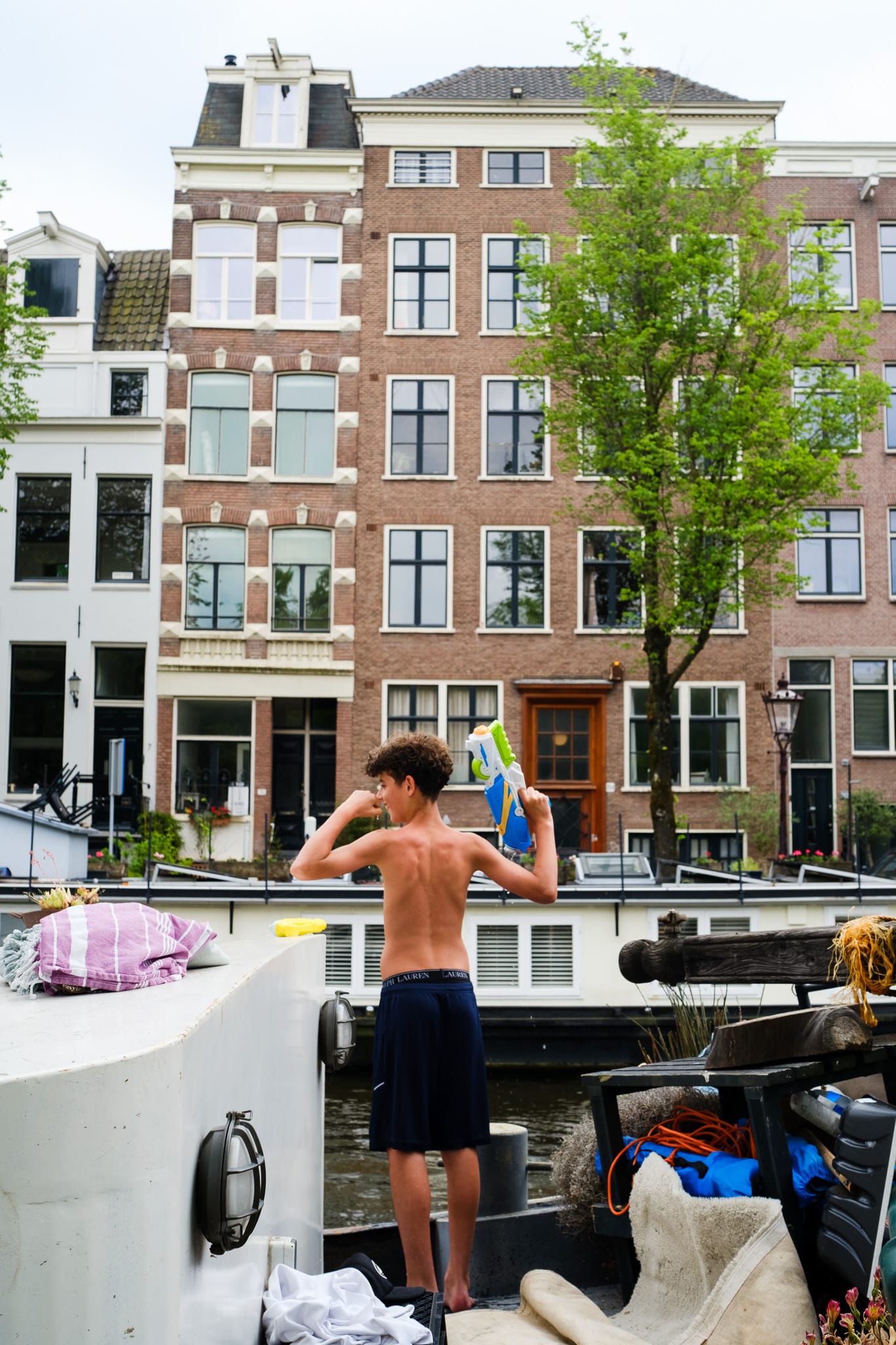 Boy standing on a houseboat on an Amsterdam canal, drying off with a towel, traditional Dutch houses in the background