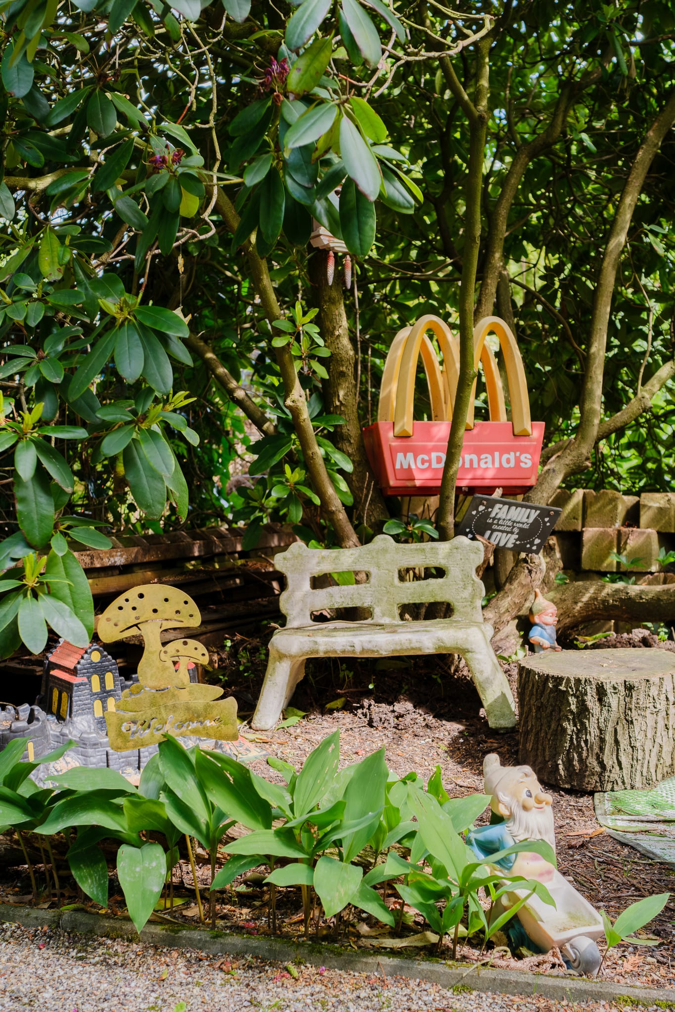 A garden corner cluttered with ornaments — a stone bench, a toy castle and a small McDonald’s sign — beneath a leafy tree