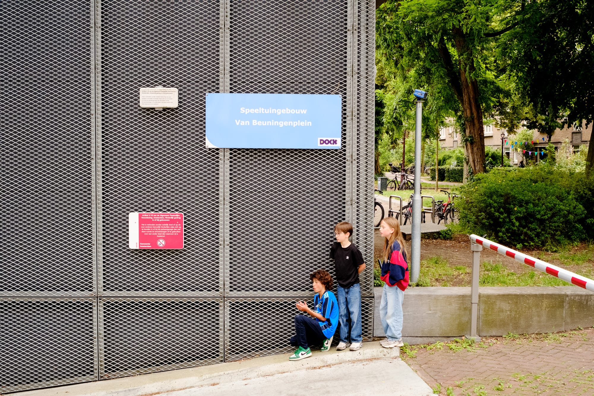 Three kids hanging out by the entrance of a playground in Amsterdam, one sitting on the ground, the others leaning against a metal gate