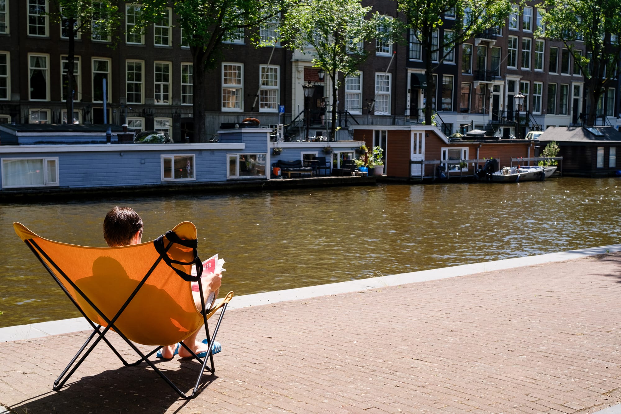 Person reading in an orange folding chair by an Amsterdam canal, houseboats and traditional houses in the background