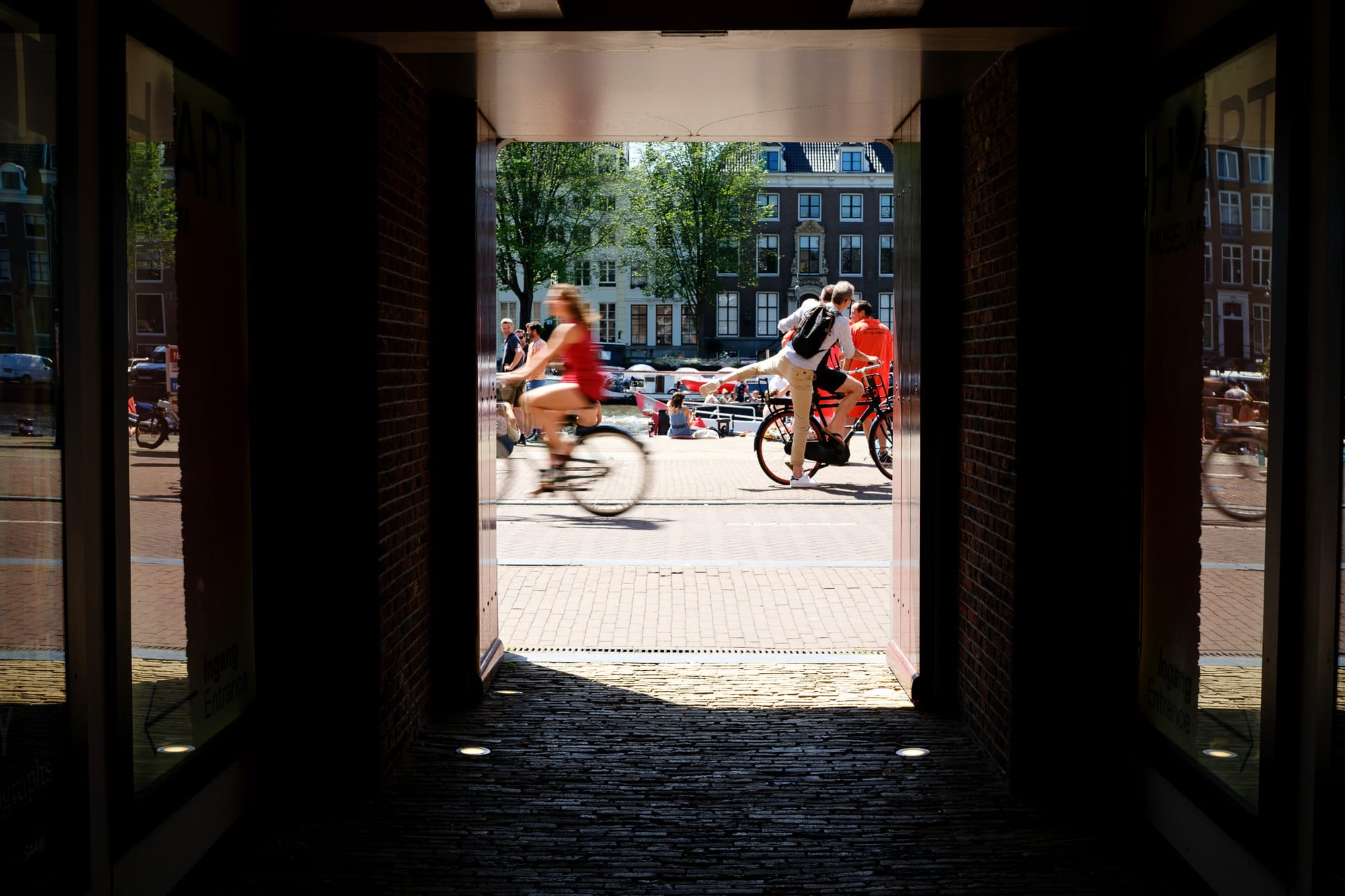 Cyclists framed through a dark archway, motion blur, Amsterdam canal and buildings visible in the bright sunlight beyond