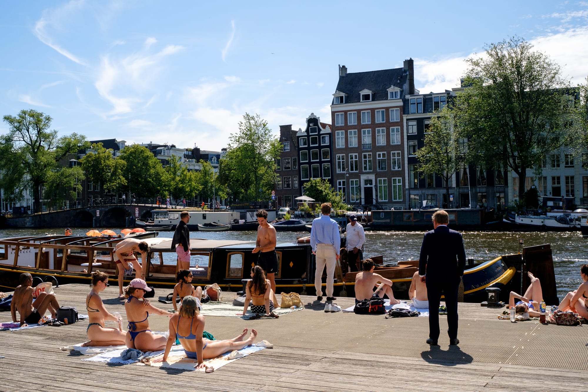 Busy Amsterdam canal-side scene with cyclists, a dog walker, and houseboats on a summer day