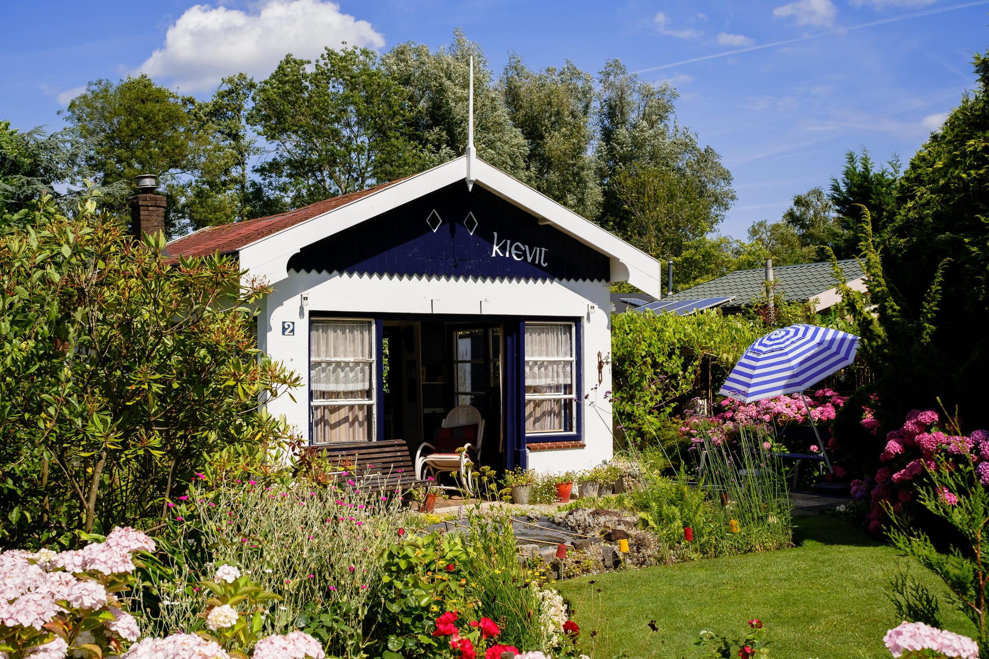 A small white and navy garden cabin named 'Kievit' with a striped awning, surrounded by a colourful flower garden and a parasol