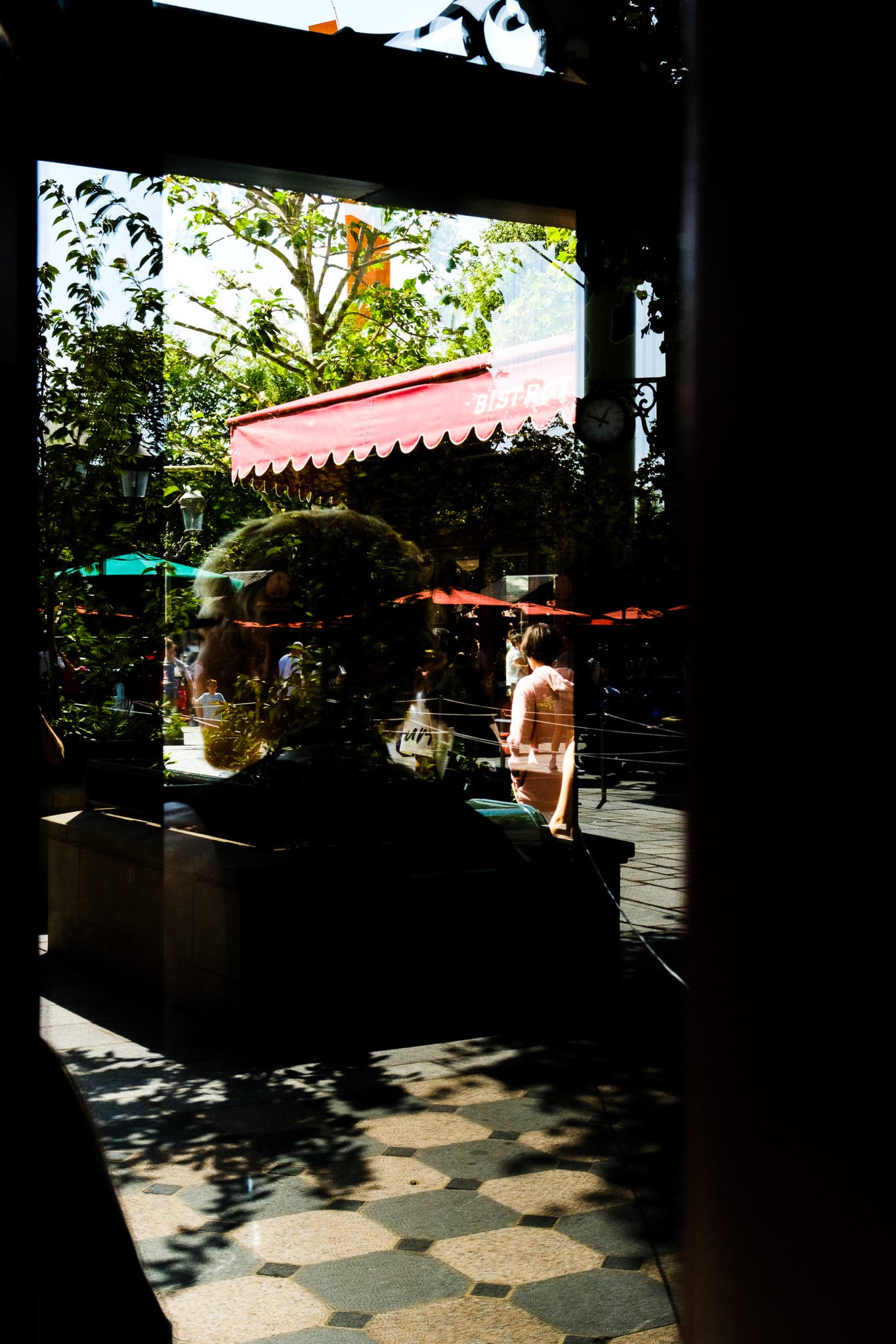 View through a dark window of a sunlit terrace with a red bistro awning, green trees, and a person in a pink shirt reflected in the glass