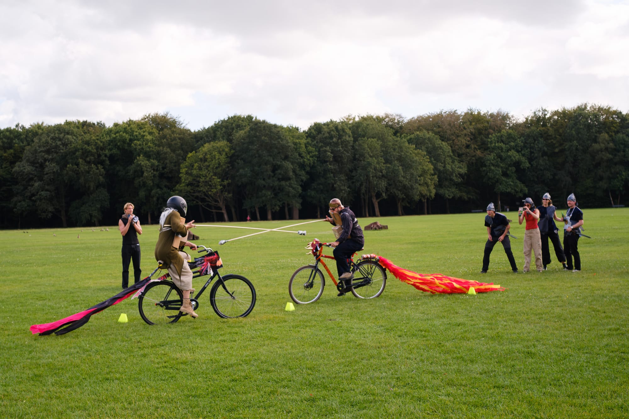 Two people on bicycles joust with long lances on a park lawn, their bikes trailing colourful streamers, while friends in homemade costumes watch