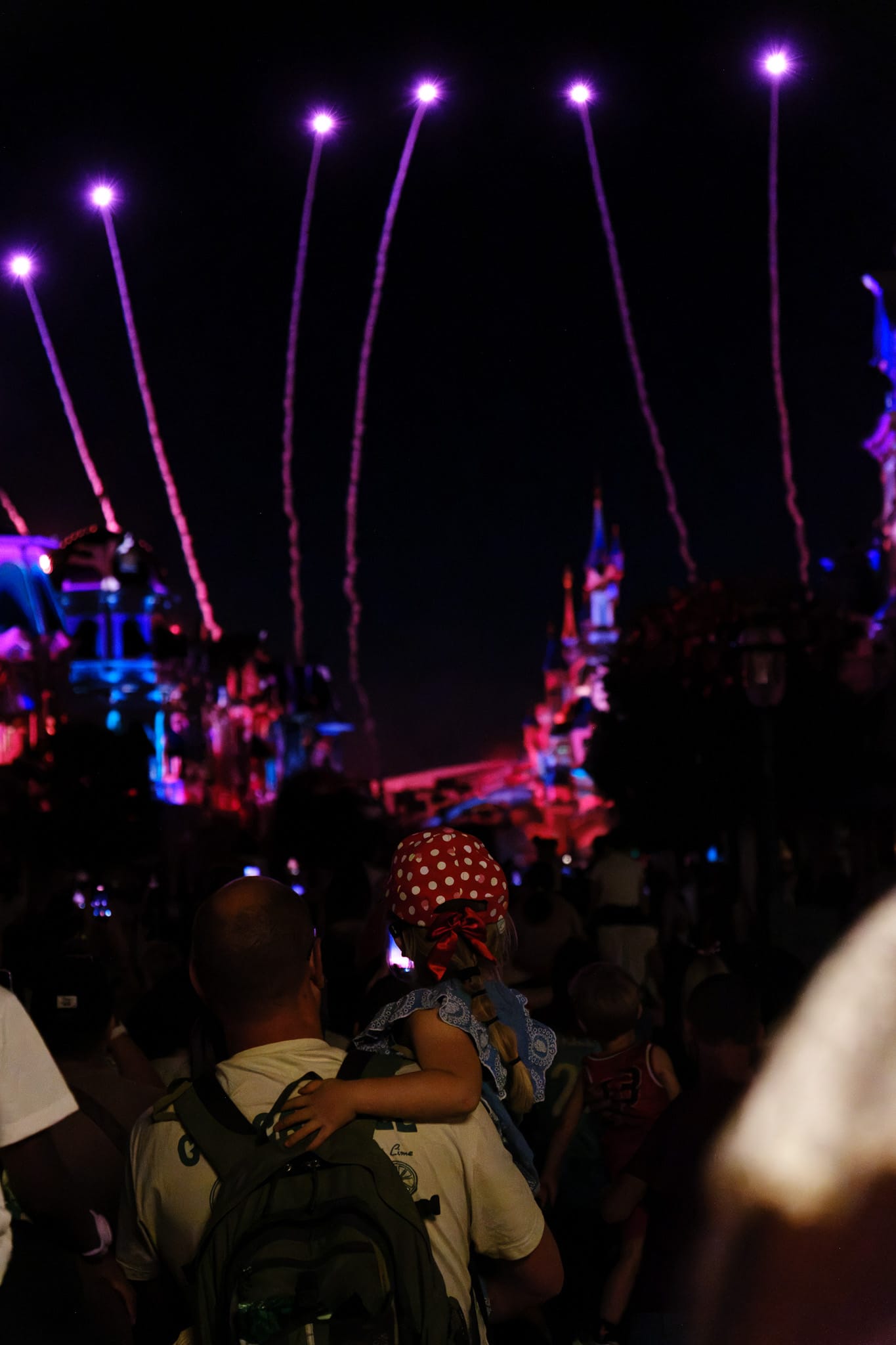 A child sitting on their father's shoulders watches pink firework trails rise into the night sky above a crowd