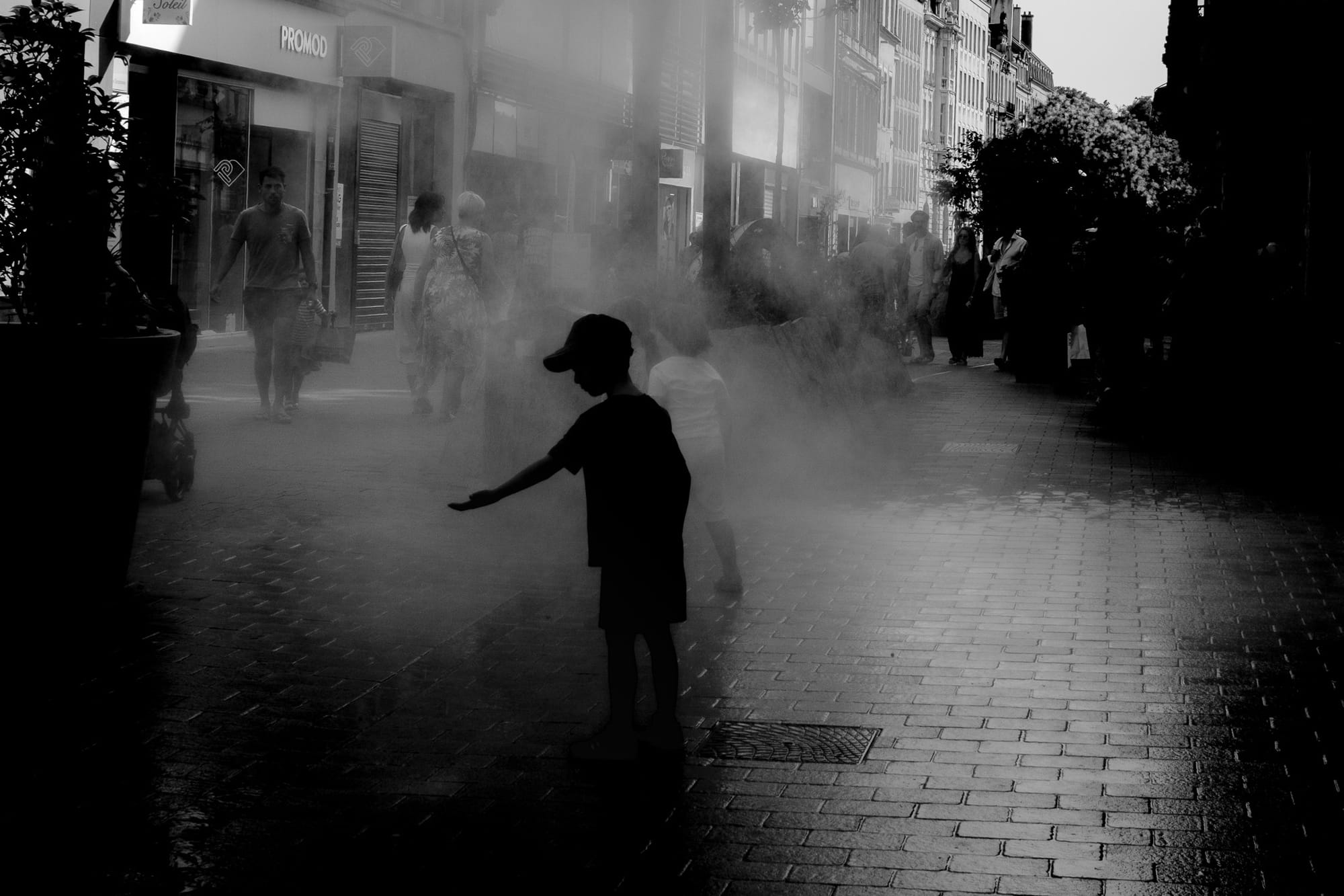 A child in a cap reaches a hand into a cloud of cooling mist on a hot shopping street, photographed in black and white