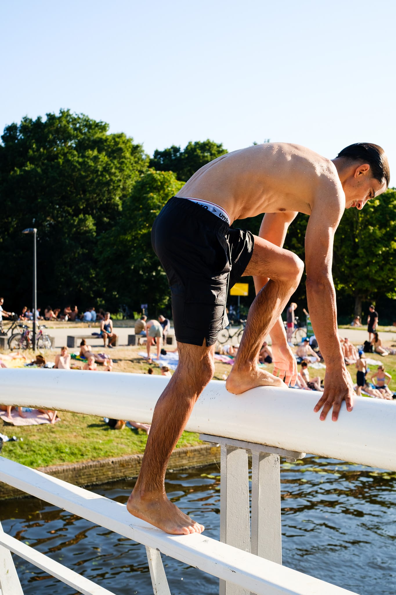 A young man in black swim shorts climbs over the white railing of a bridge to jump into the canal, with people sunbathing on the grass behind
