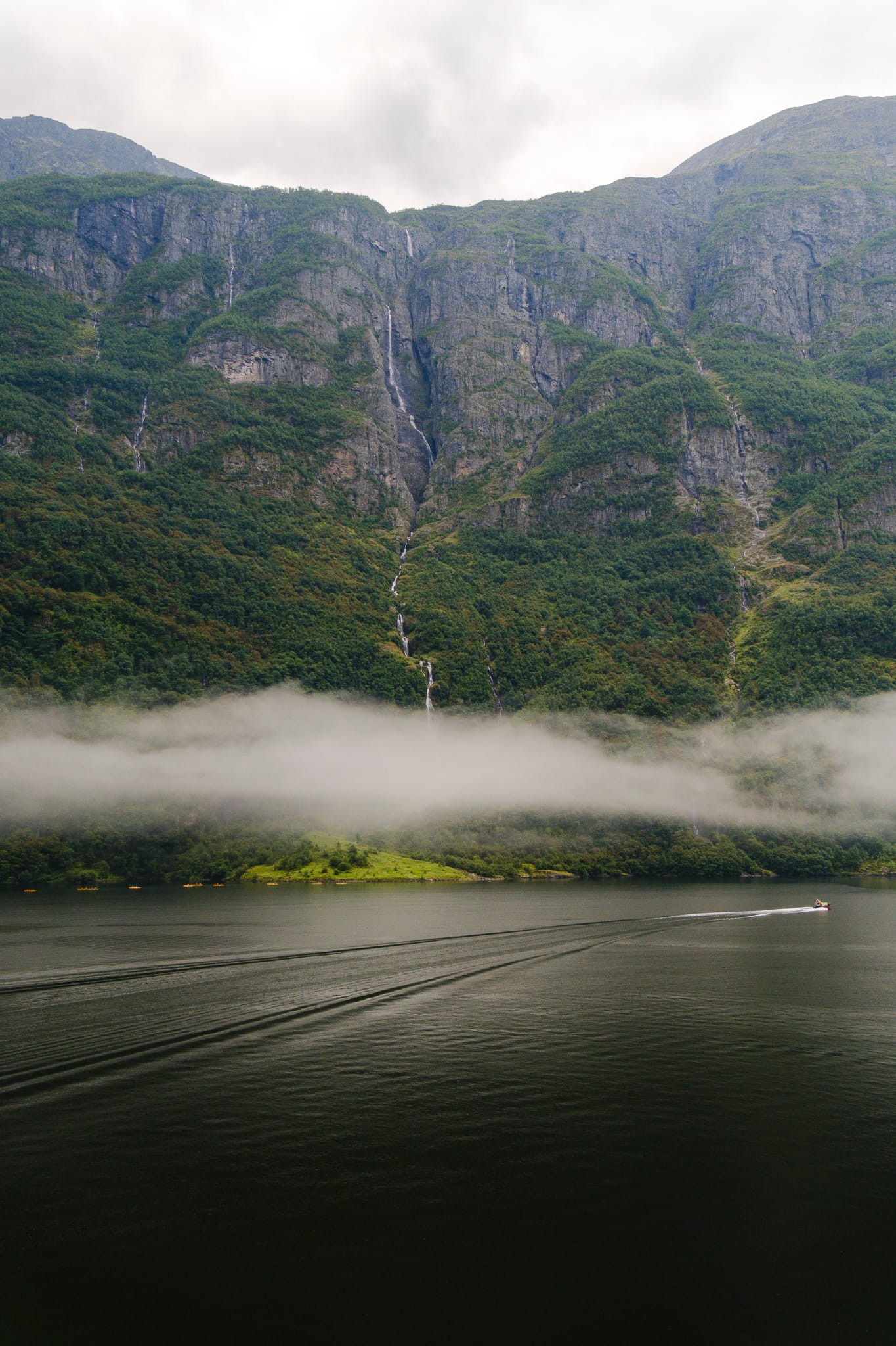 A thin waterfall running down a green cliff into the fjord, with a band of low cloud over the dark water.