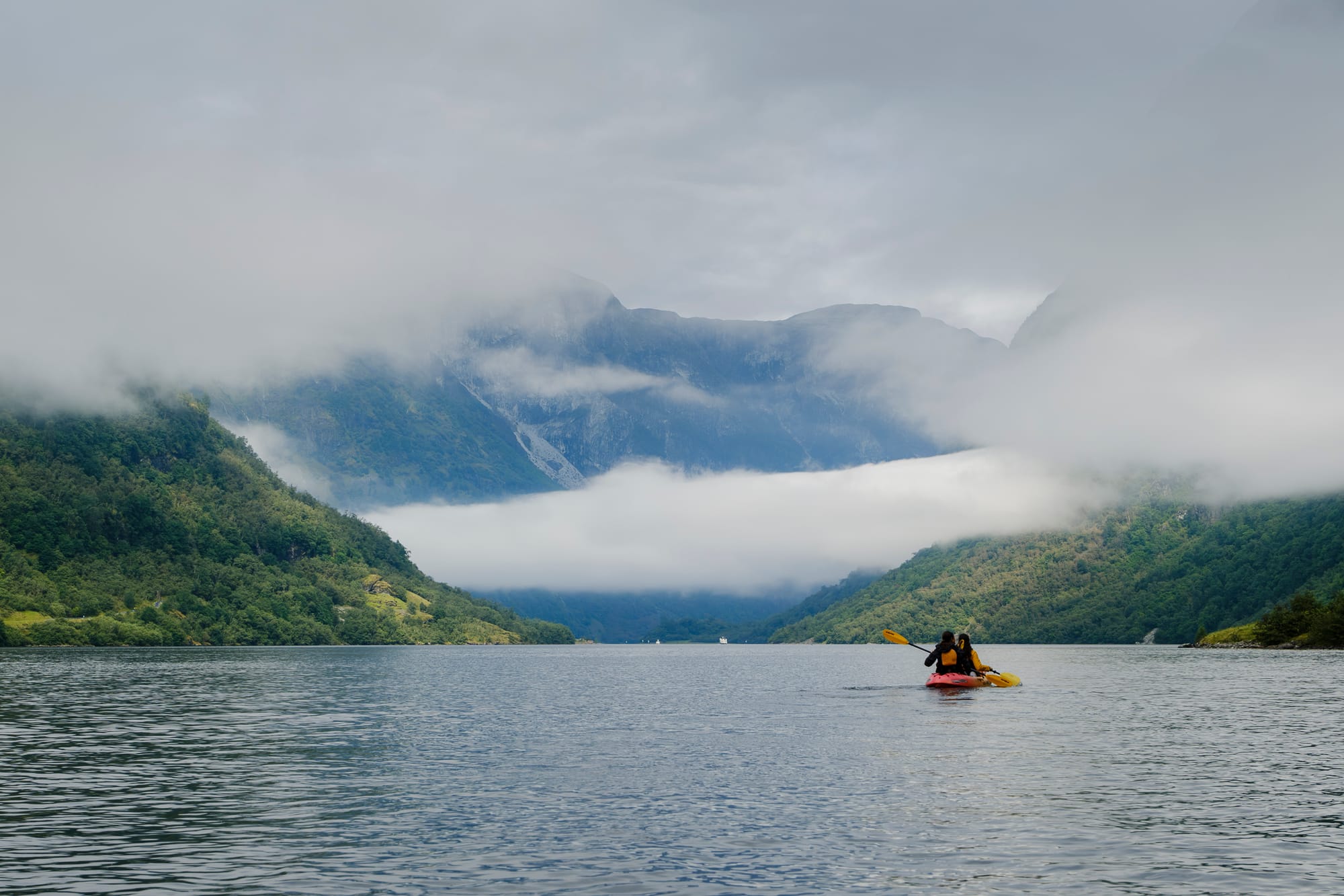 Two people in a red kayak on the fjord, with low cloud covering the mountains behind.