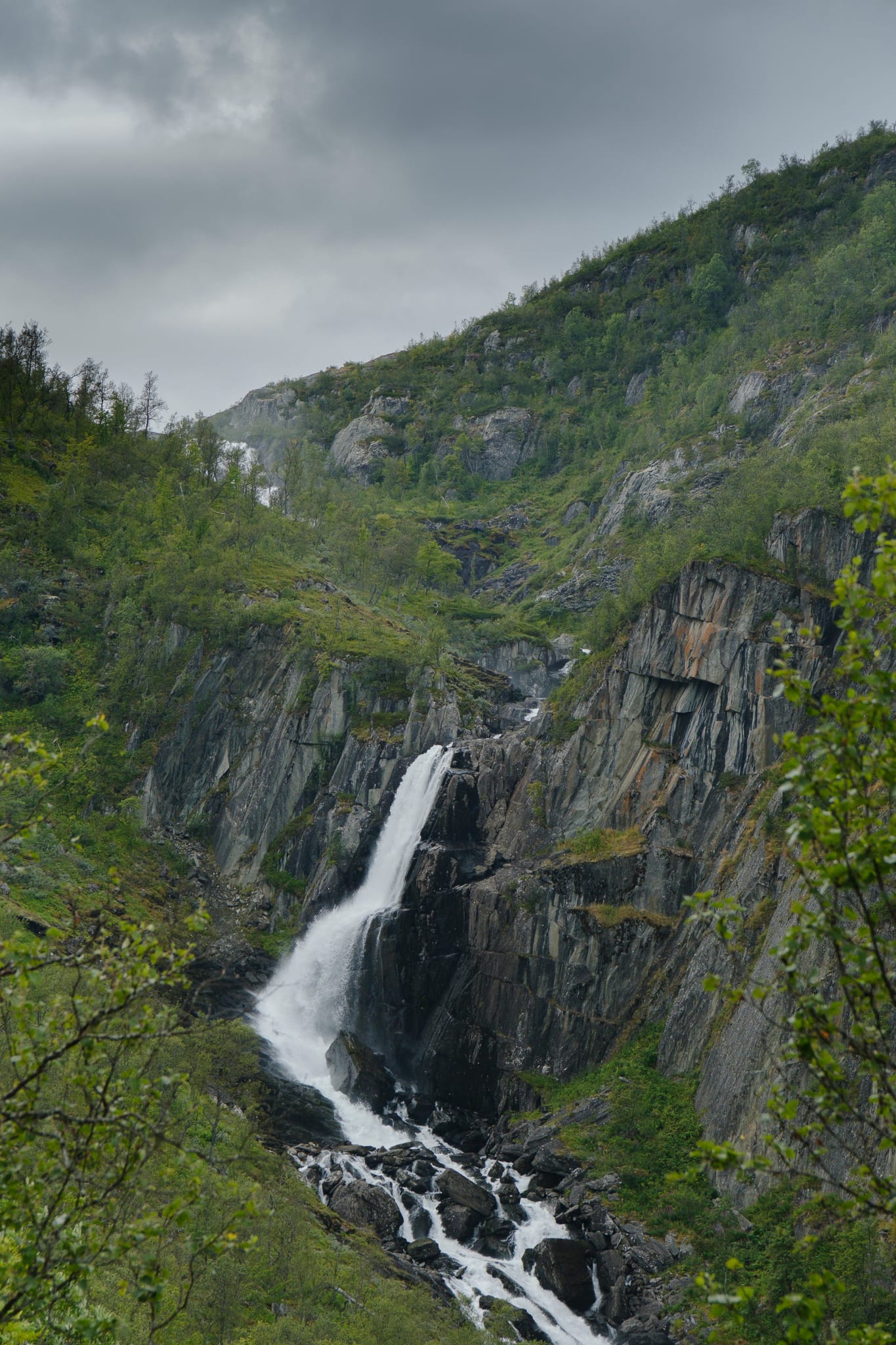A waterfall dropping down a dark rock face into a narrow green gorge.