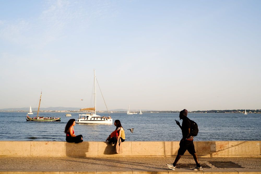 Three people by the Lisbon waterfront at golden hour, two sitting on a stone wall with sailboats on the Tagus river, one walking past checking their phone