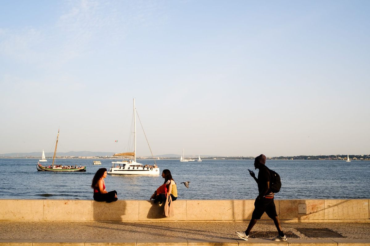 Three people by the Lisbon waterfront at golden hour, two sitting on a stone wall with sailboats on the Tagus river, one walking past checking their phone