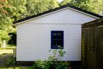 A white timber garden cabin with a blue-framed window and a wooden door, set among green trees and shrubs