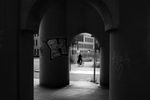 A cyclist passes behind the arched concrete columns of a bridge underpass covered in graffiti, photographed in black and white