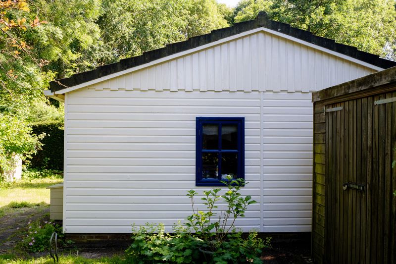 A white timber garden cabin with a blue-framed window and a wooden door, set among green trees and shrubs