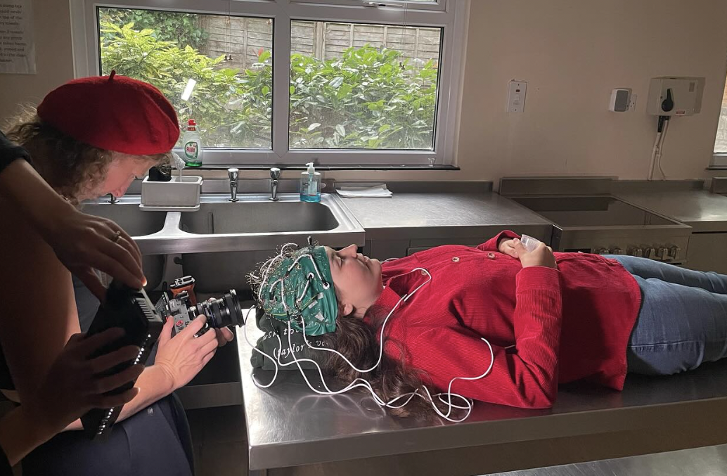 A person is lying on a metal table with a hat covered in wires, whilst another person is filming them.