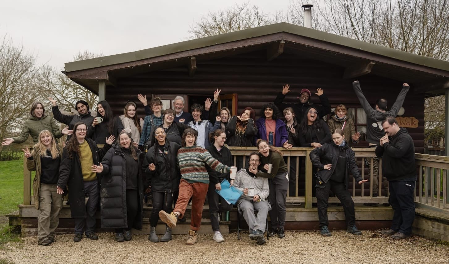 Crew of 26 posing for a silly photo in front of a rustic cabin