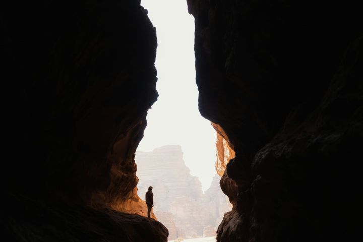 A lone person stands inside a dark canyon cave, looking out towards bright desert cliffs beyond.