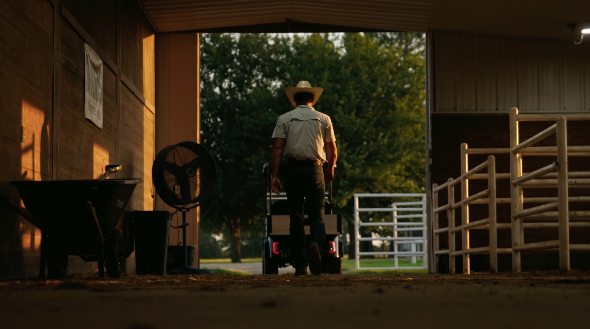 Silhouetted against the daylight, a rancher in a cowboy hat walks out of a dark barn toward a Denago OXEN parked in the doorway