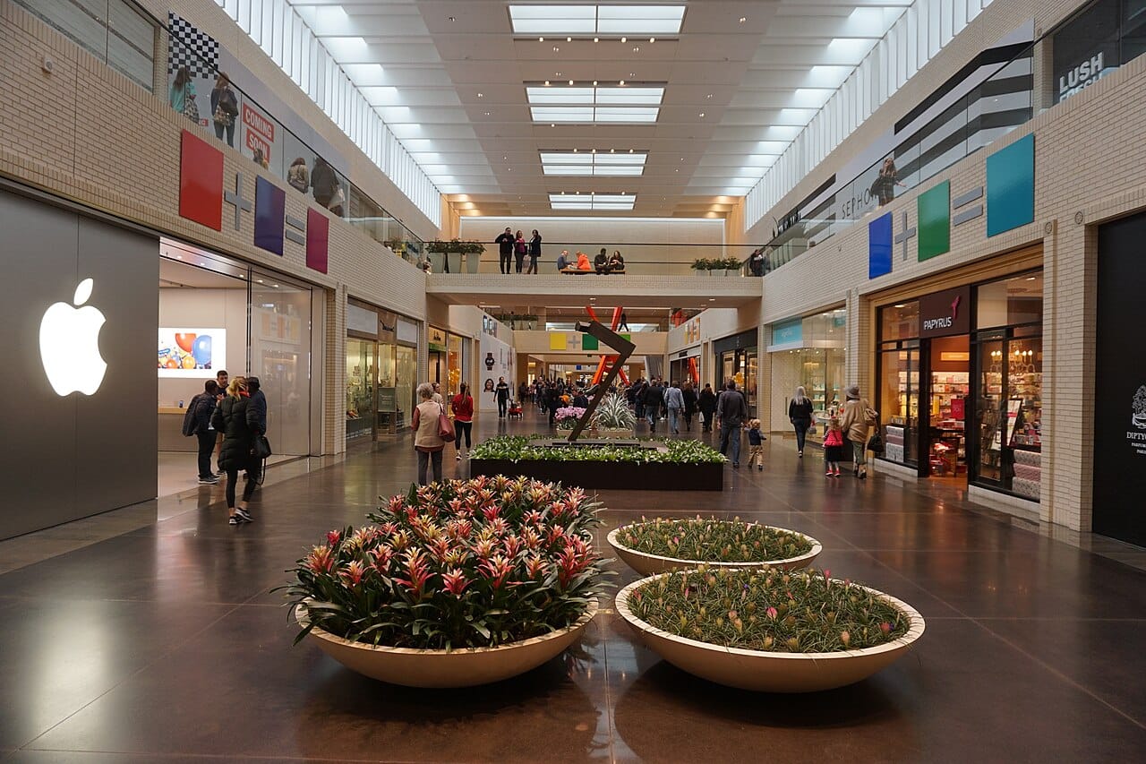 The concourse at NorthPark Center by the Apple store, with an Anthony Caro rusted steel sculpture in the middle distance, the orange steel of Ad Astra beyond it, and Color Equations panels on both walls.