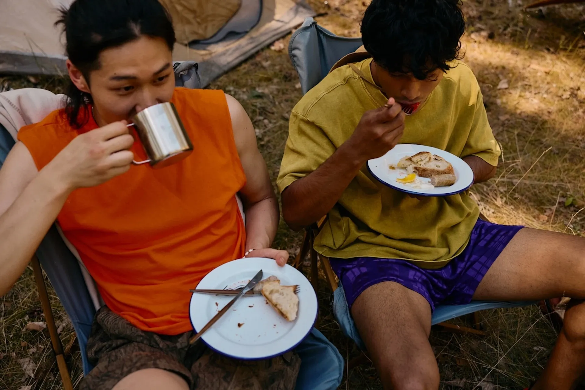Two friends in camping chairs, drinking coffee and eating sandwiches