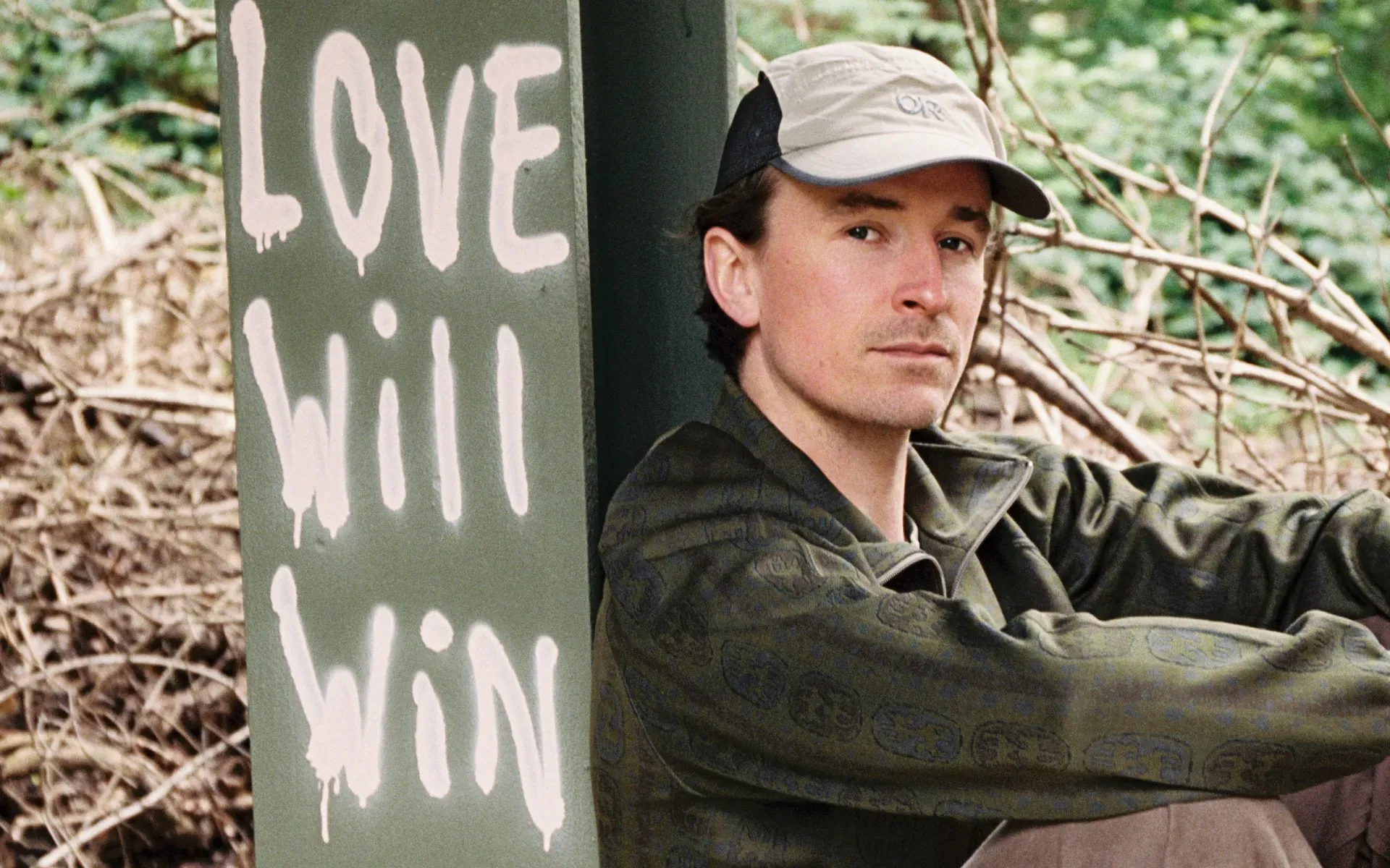 Ross From Friends sitting next to some graffiti, which says “Love Will Win”.
