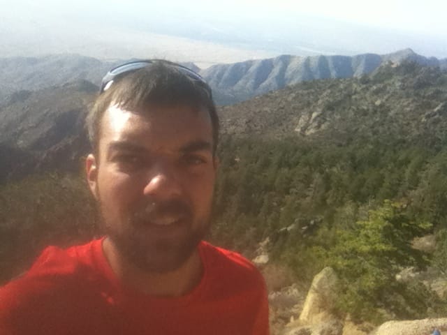 Selfie of man in a red shirt. Sunglasses are on the top of his head. Mountains are behind him.
