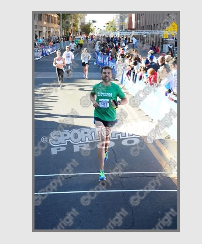 A man in a green shirt and short running shorts running across the finish line of a race.