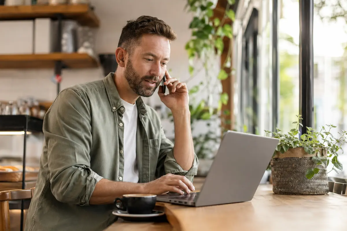 A small business owner reviewing website enquiries from a bright café while talking on the phone, with no visible text or logos