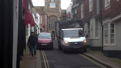 A car is forced up on the pavement in order to pass a parked lorry, and the pedestrians are unaware of the danger behind them.