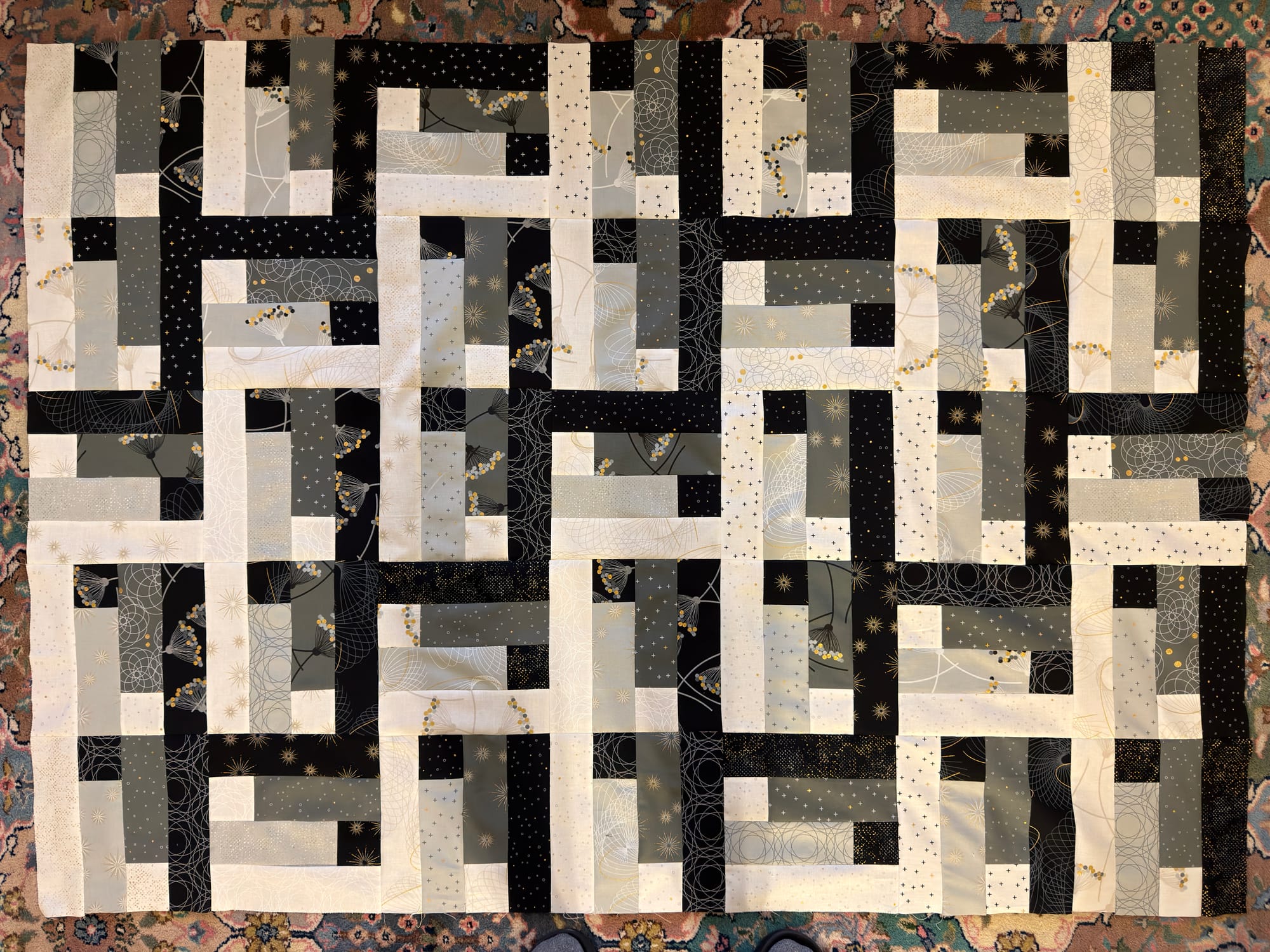 Quilt top with black white and two tones of gray patterned fabric, creating a zigzag stare pattern that almost looks woven like a herringbone. The block pattern repeats with block, block, reflected Blox rotated 90°. The layout is a 7 x 5 pattern. The block has a dominant black strip on the right and white strip on the left with an eye then an exclamation between made of jelly roll fabric strips.