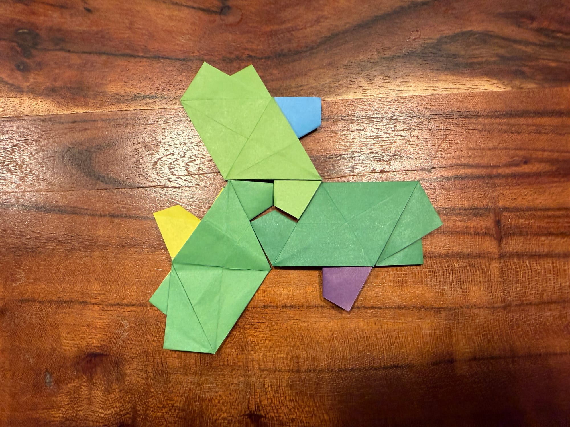 three aperiodic turtle origami peices with all three heads in the center forming a triangle. the tiles are all green with one leg a different color. one is blue, one is purple, and one is yellow. they are on a wooden table. 