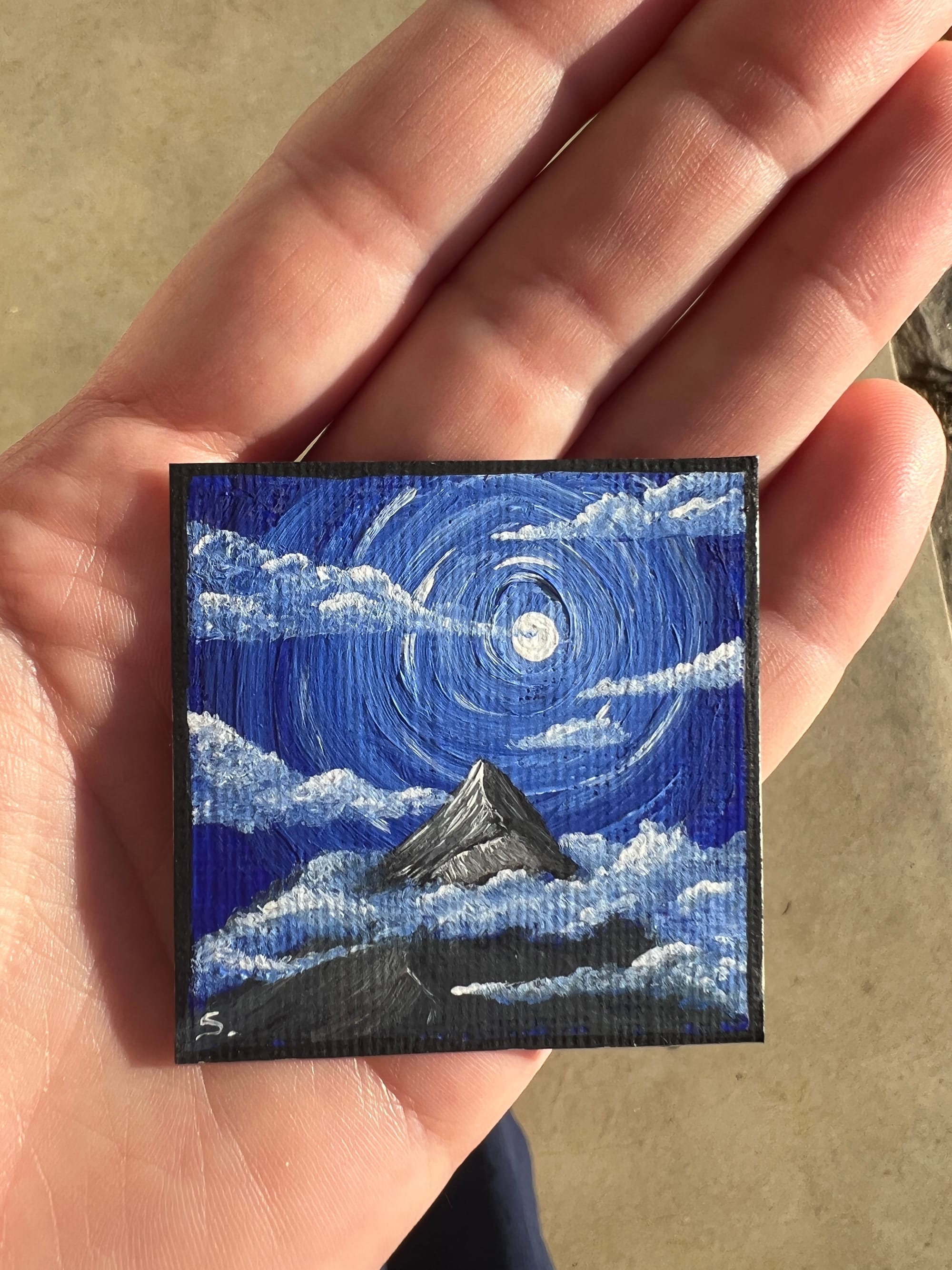 a hand holding a small 2in painting of a mountain with clouds  with a moon and blue sky.