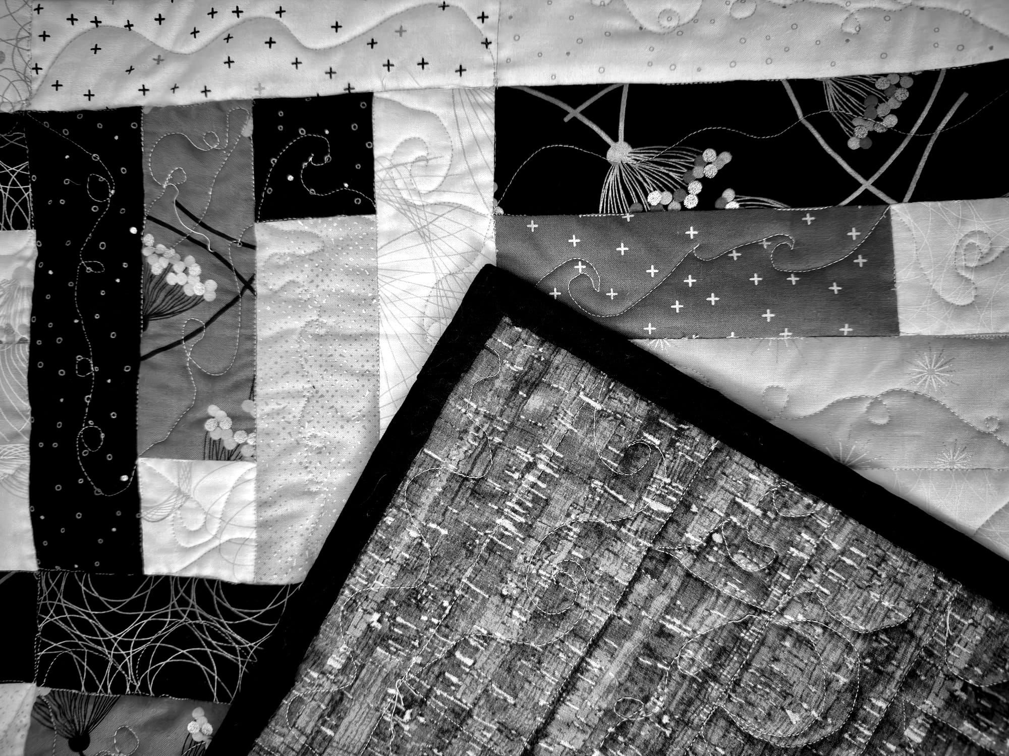 The corner of the back of a quilt on top of a jellyroll pattern of black, white, and grey fabrics with flowers, plus, stars, and circle patterns. 