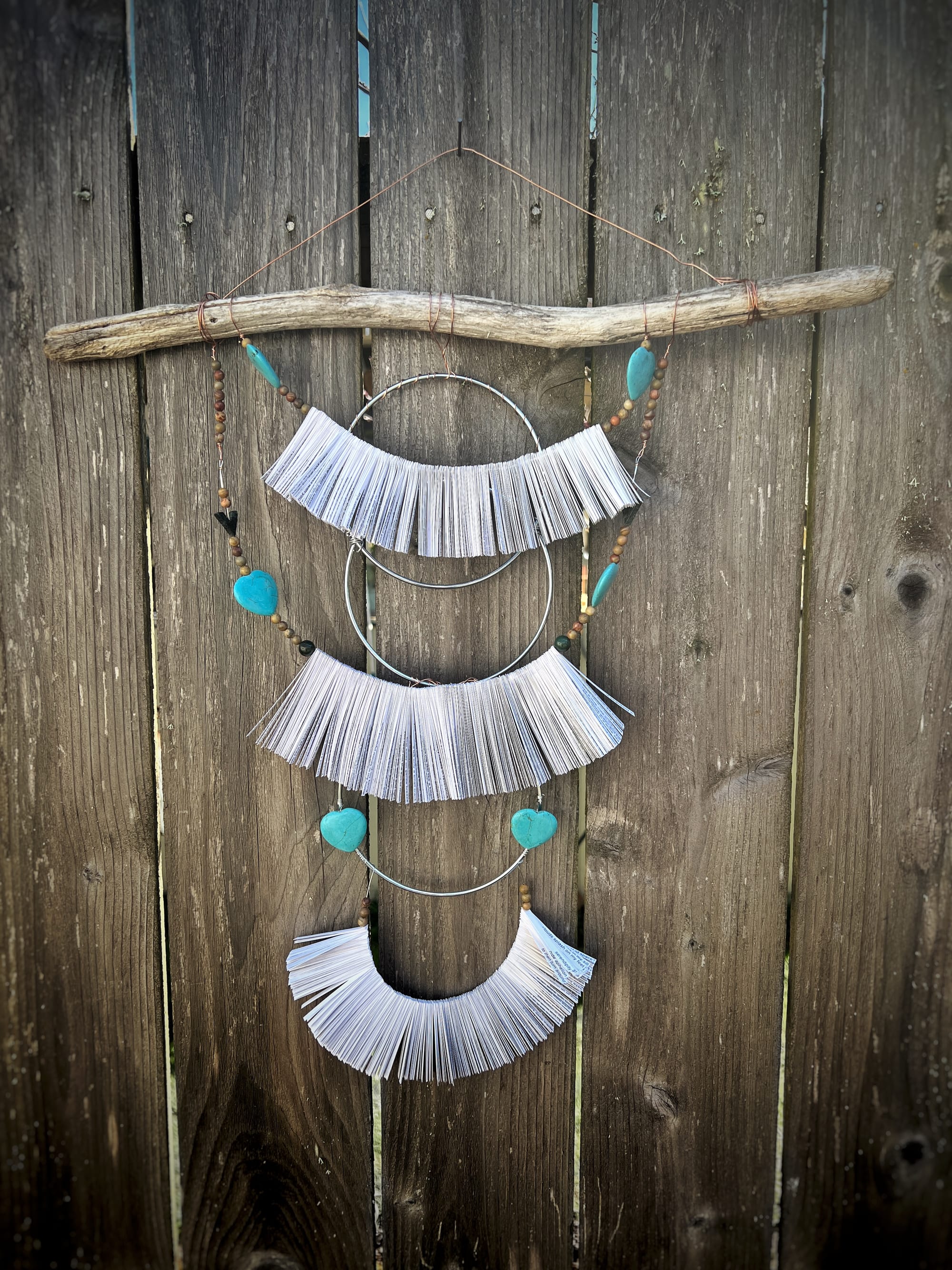 a work of art with a stick with 3 draping wires full of paper strips and beads with circle hoops incorporated hanging on a fence