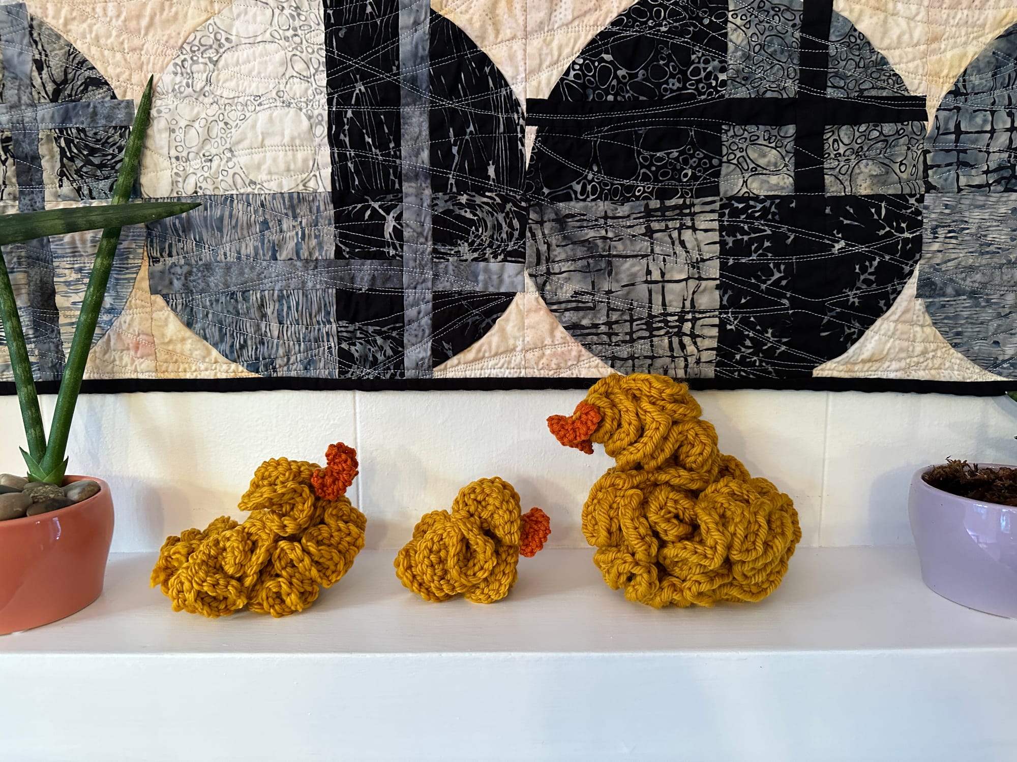 3 hyperbolic yarn ducks on a mantle with a quilt in the back ground and plants on the sides. 