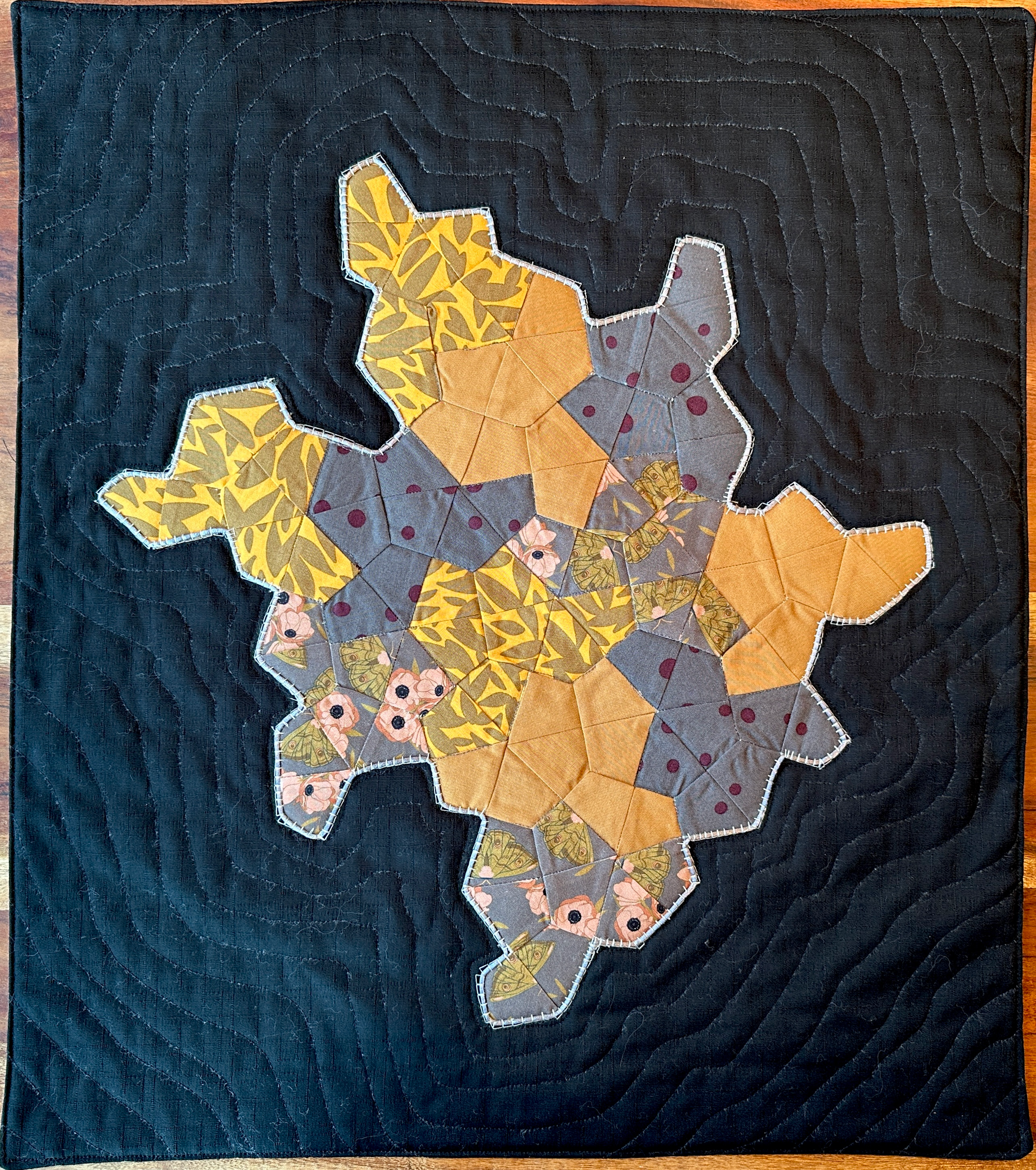 Image of monotile quilt. 12 tiles place together with 4 different materials on a black background. materials are grey polkadot, grey with butterfly, solid tan, and yellow with leaves