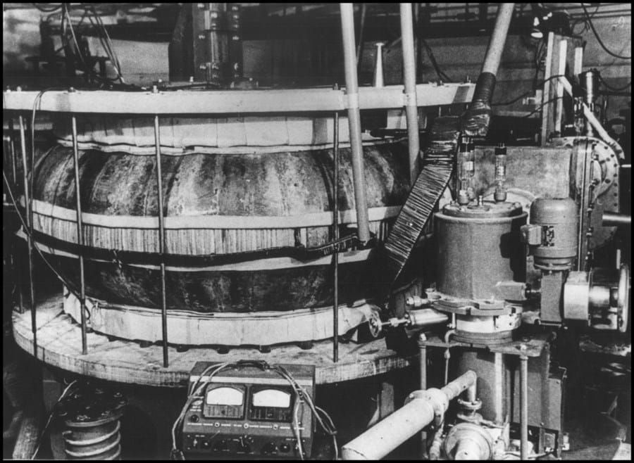 A black and white photo of a tokamak, a donut-shaped device surrounded by various piping and other industrial and electrical machinery.