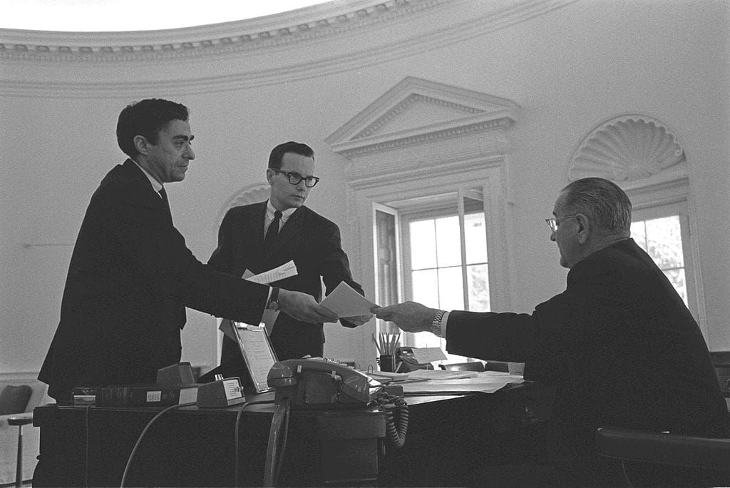 Staffers hand Lyndon Johnson documents inside the Oval Office