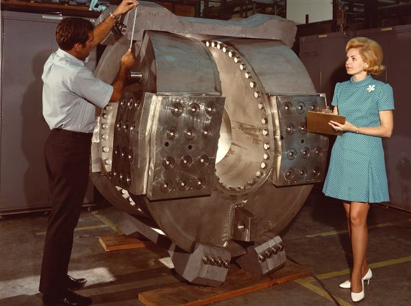A man and a woman stand next to a large metal structure used in a fusion experiment, making measurements, in the 1960s