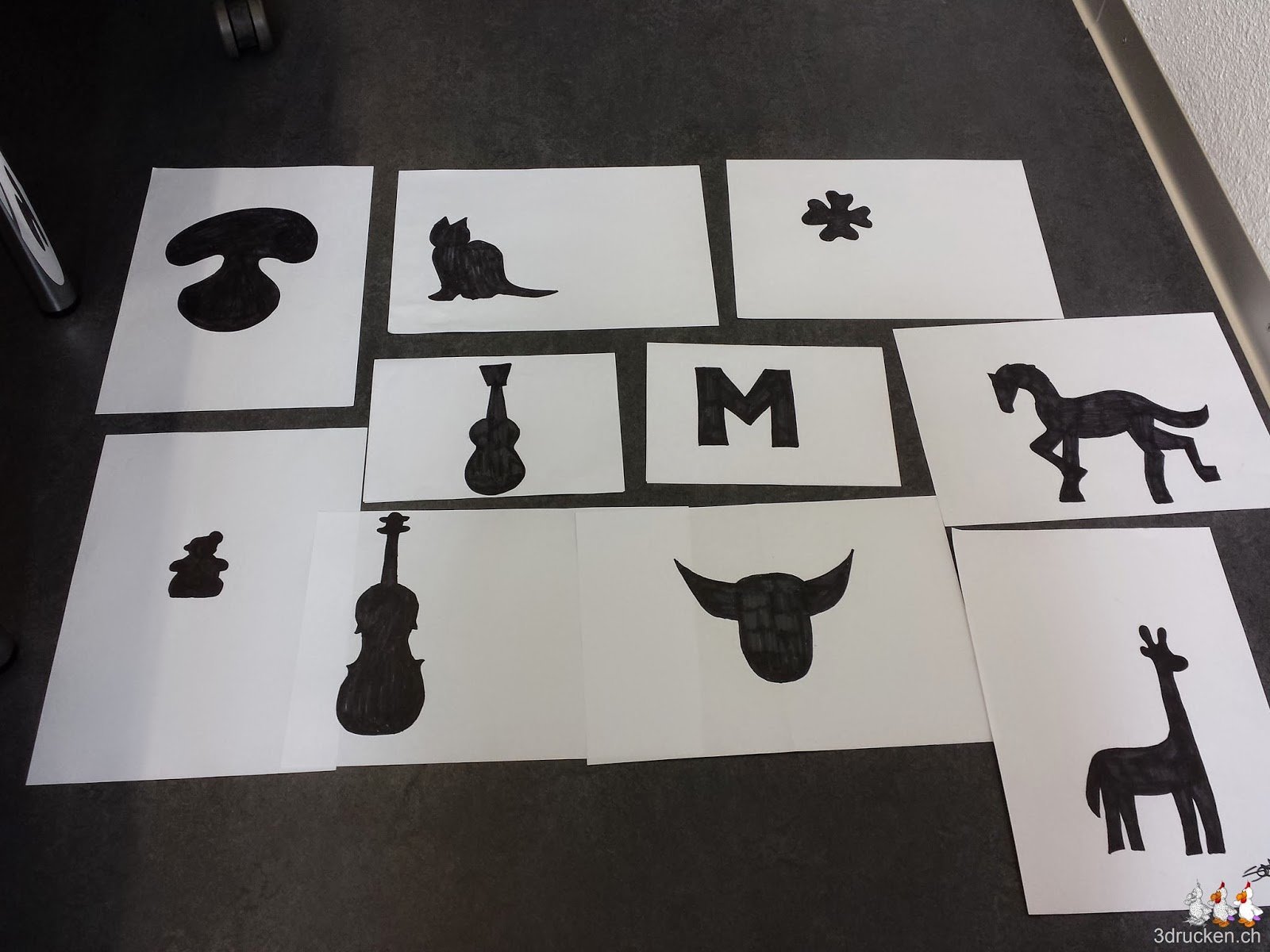 Foto der am Boden ausgelegten Handskizzen mit schwarzem Filzstift ausgemalt. Tiere, Natur, Musik und ein M