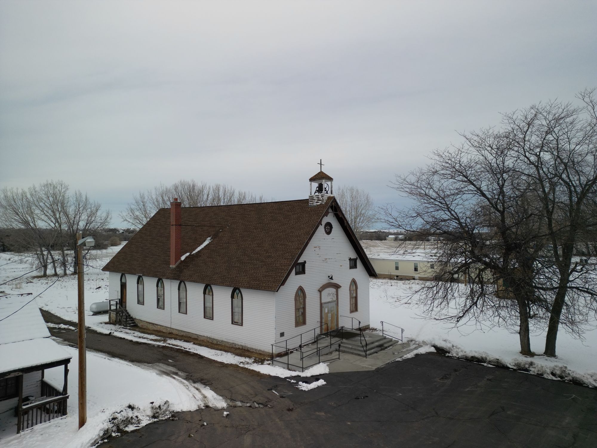 An aerial photo of St. Francis Xavier Mission Church in Montana in the winter. The Church is a simple painted wood frame clapboard structure with gothic windows and a simple gable roof with a small cupola.. 
