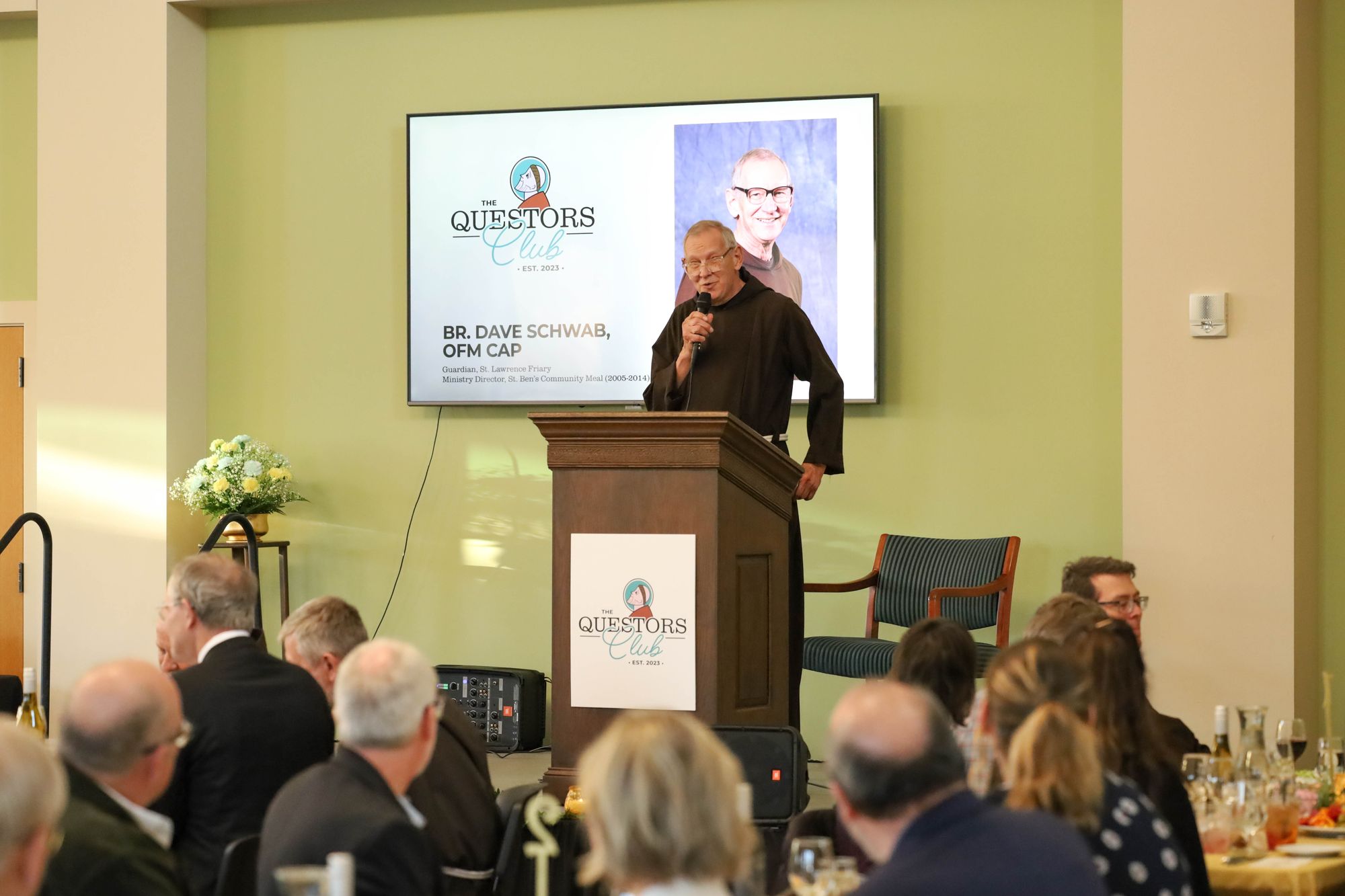 Color photograph of Br. Dave Schwab, OFM Cap. at the podium giving the blessing before dinner.