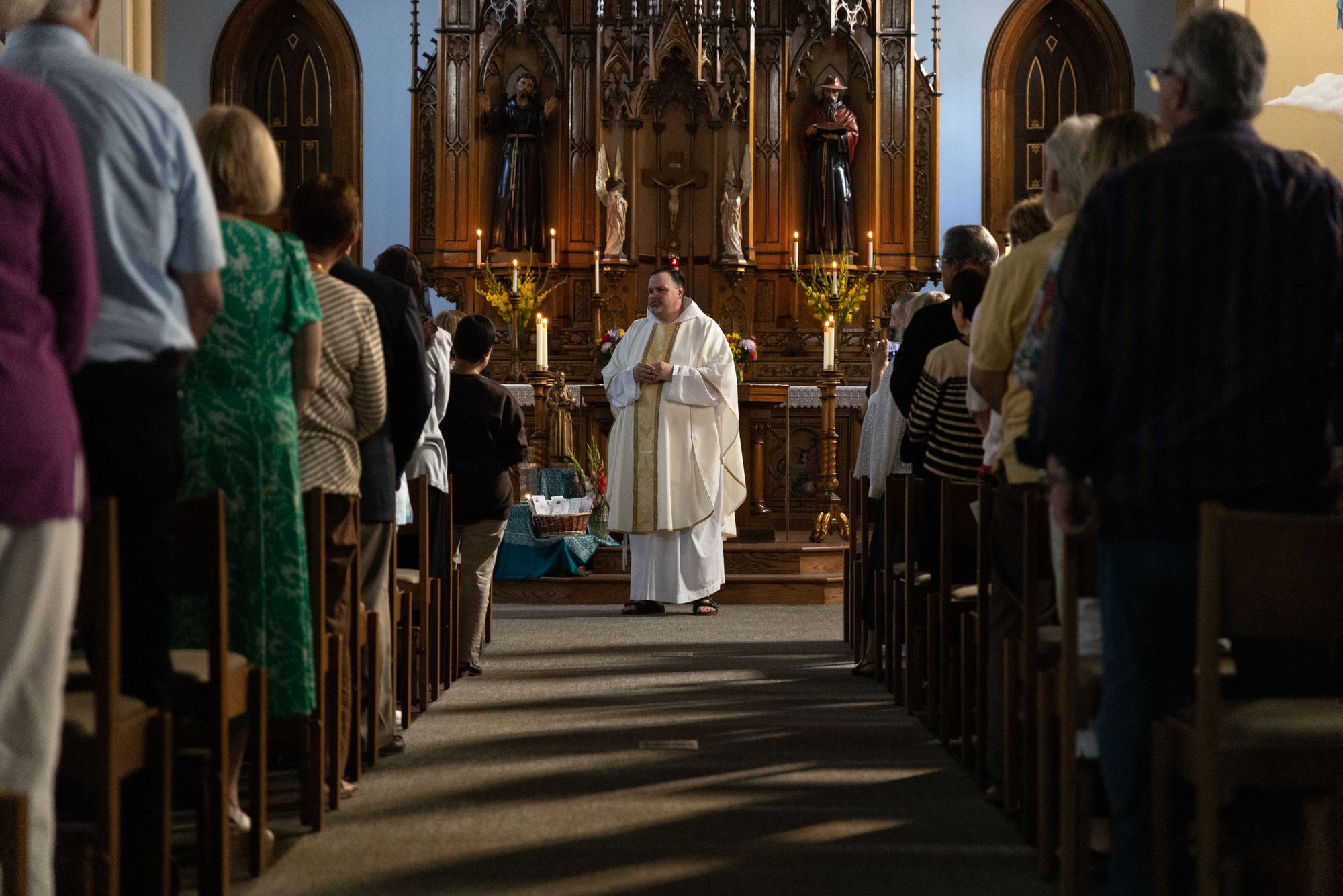 A color landscape photo looking up the center aisle of St.Bonaventure Chapel toward the sanctuary, where Br. Steve Kropp, OFM Cap. is preaching the Feast Day homily on the Feast of Blessed Solanus Casey on July 30, 2023. The artificial illumination is off due to a power outage and there are candles lit at the altar.