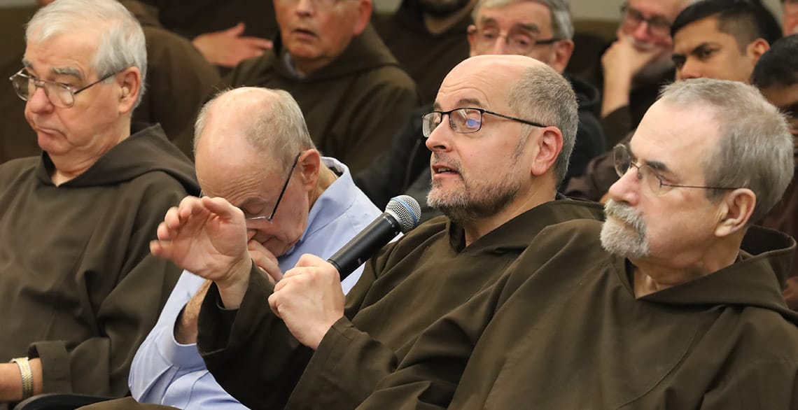 Br. Robert Wotypka is seated among a group of Capuchin friars in an auditorium. He is holding a microphone and speaking.