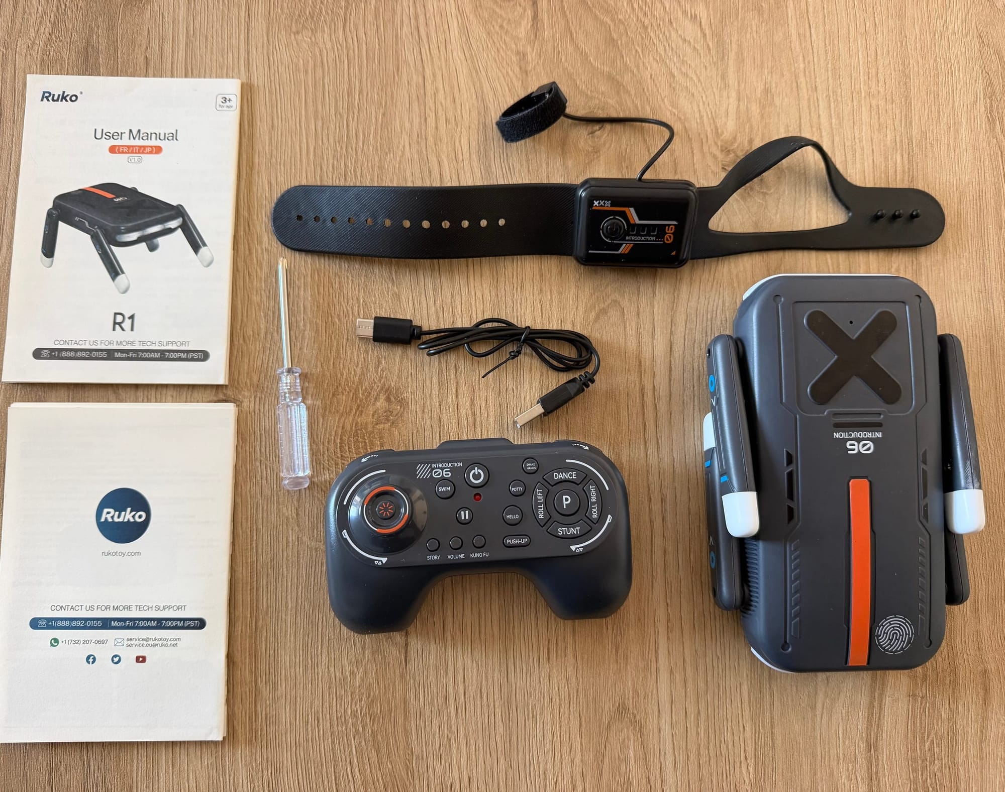 Ruko R1 robot, handheld remote, gesture watch, USB cable and printed documents laid out on a wooden table.