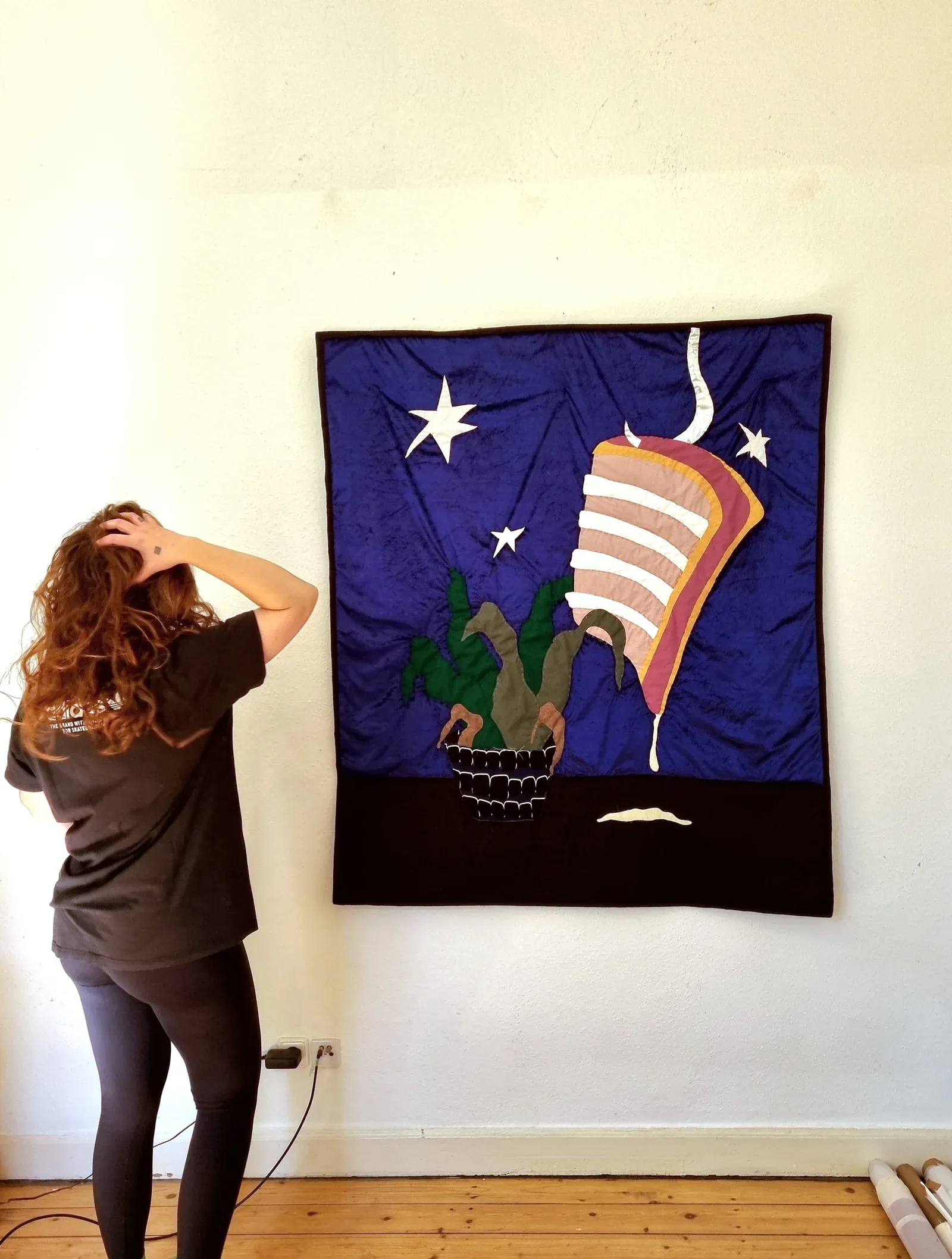Rosina Rosinski standing in front of her 2025 textile artwork Rippe bei Nacht II, made from cotton, velvet, and polyester. The piece features a stylized nighttime interior with smoke, stars, and plants, evoking solitude and emotional distance.