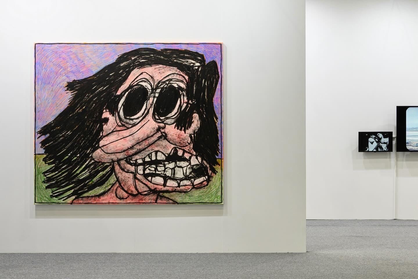 Artwork by Taylor A. White titled Dawn, Wind (2023), exhibited at Taipei Dangdai. The painting shows a large, distorted cartoon-like figure with black hair, wide oval eyes, and exaggerated teeth, drawn in thick black lines over pink, green, and multicolored textured backgrounds. The work is mounted on a white exhibition wall in a fair setting, with video works visible in the adjacent booth.